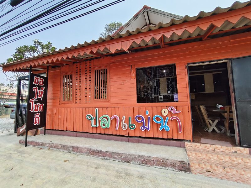หน้าร้านบ้านไม้สีส้ม มีตัวอักษรหลากสี ปลาแม่น้ำ และป้าย ขอให้โชคดี